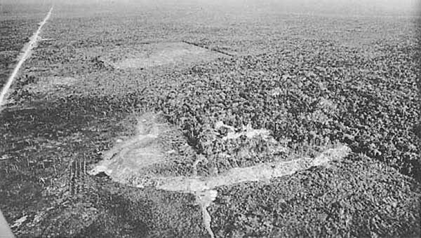 Aerial photograph of Becán in the Yucatán jungle. The defensive moat is visible as a continuous cleared band encircling the central pyramid complex, distinguishing the fortified core from the surrounding forest.