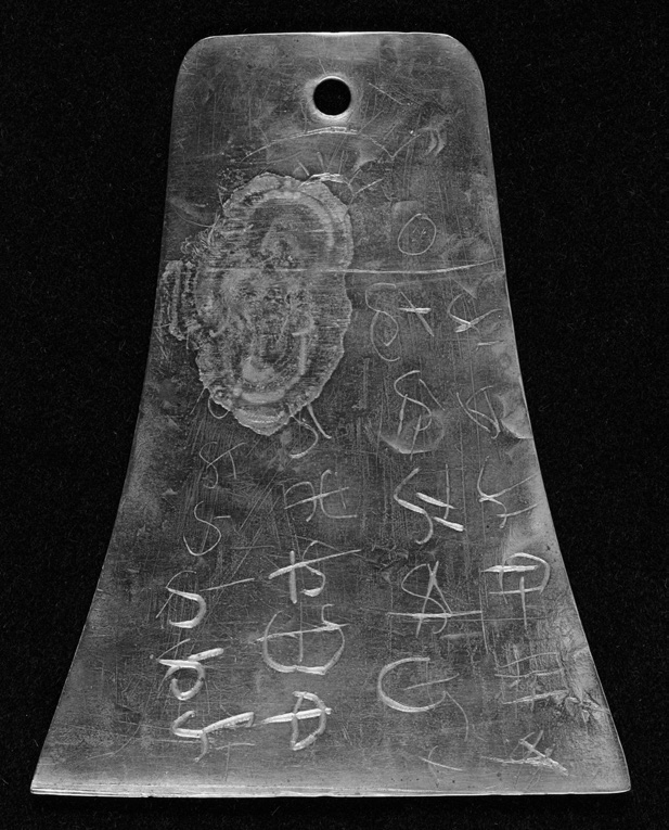 Photograph of the surviving Kinderhook plate held by the Chicago History Museum, a small, dark, bell-shaped brass plate roughly four to five inches long, incised on both faces with rows of crude hieroglyph-like characters arranged in horizontal registers.