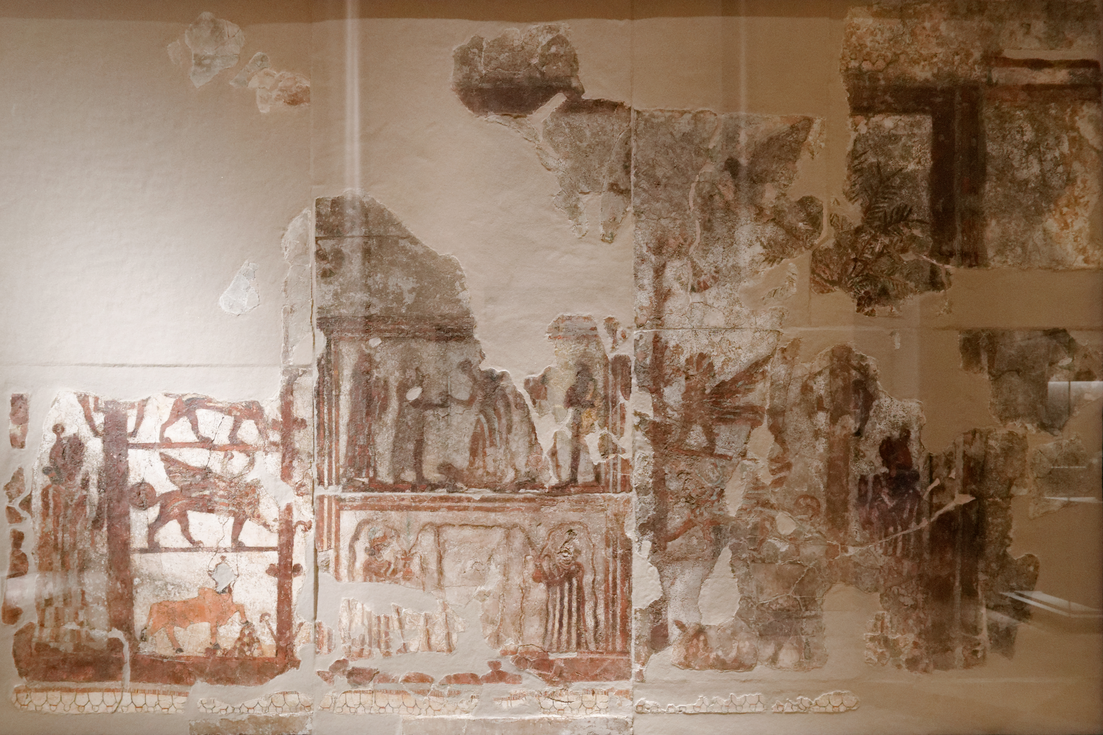 Color photograph of the Mari Investiture Panel — an Old Babylonian tempera-on-plaster wall painting circa 1800 BC, excavated from the Royal Palace of Mari in the 1930s. The panel shows King Zimri-Lim receiving the rod and ring of authority from the goddess Ishtar in a temple setting, flanked by lesser deities, guardian figures, and stylized palm trees representing a sacred garden.